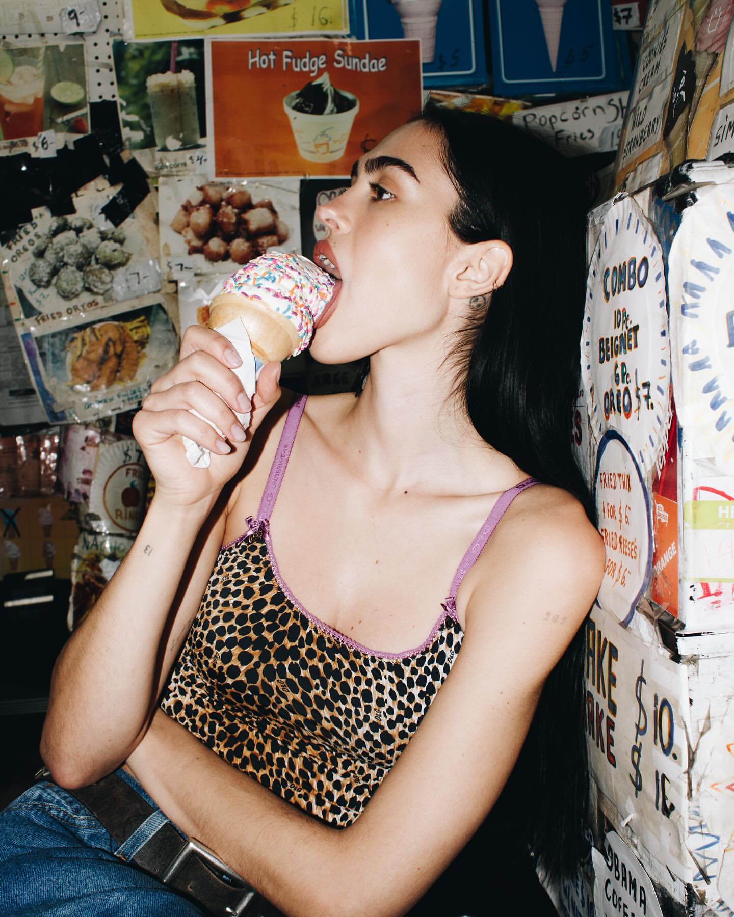 Young woman in a leopard print top is eating an ice cream cone in front of a wall covered with food advertisements in an indoor setting.