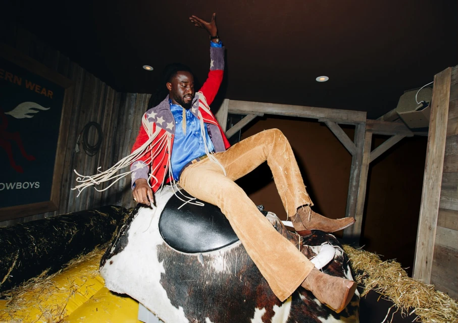 An adult male wearing a colorful fringe jacket rides a mechanical bull in an indoor venue with dim lighting and rustic Western-themed decor.