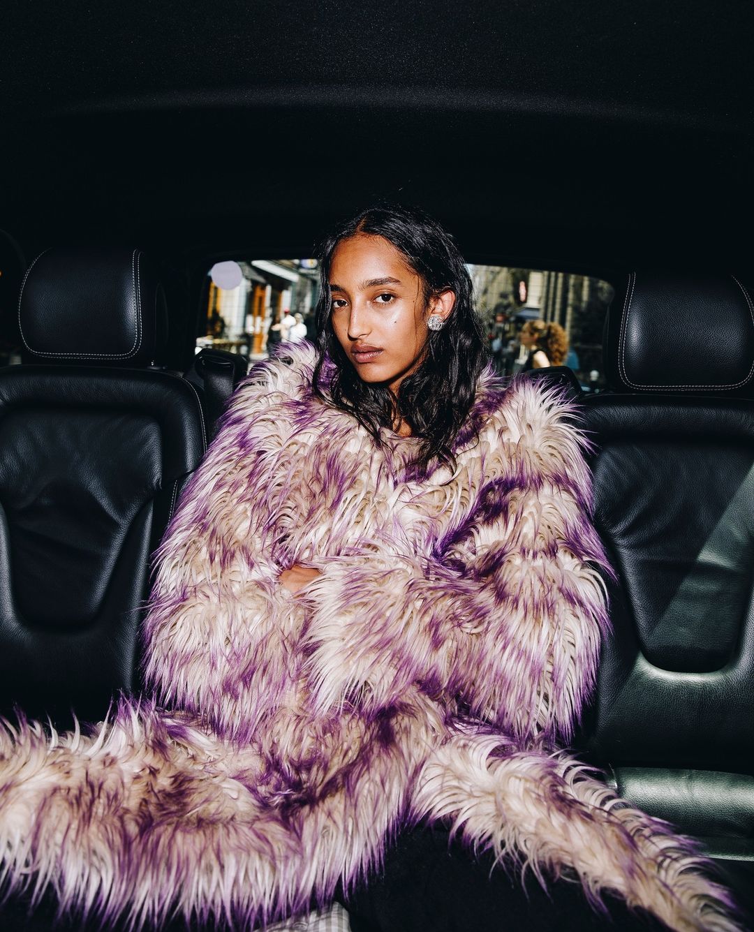An adult female sits in a car wearing a lilac and white faux-fur coat, looking at the camera with a neutral expression and dark, shoulder-length hair.