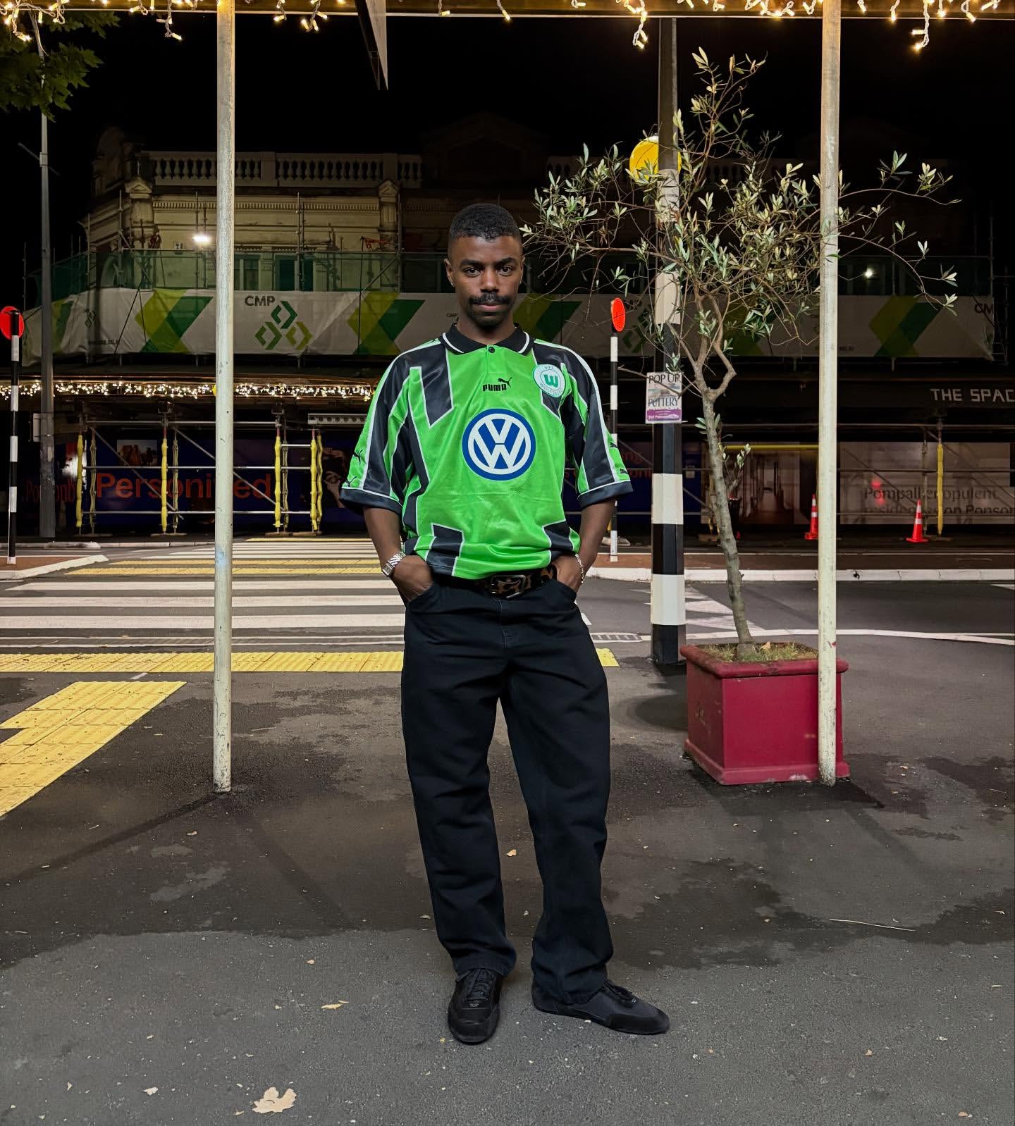 Adult male in streetwear fashion stands on a crosswalk at night, wearing a green soccer jersey and black pants, lit by streetlights.