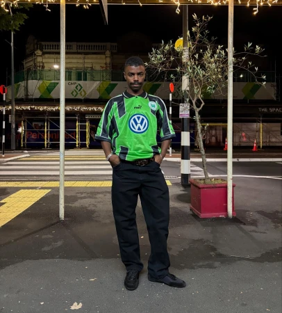Adult male in streetwear fashion stands on a crosswalk at night, wearing a green soccer jersey and black pants, lit by streetlights.