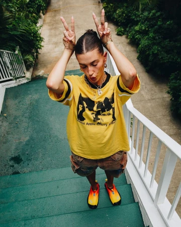 Adult female poses on blue stairs outdoors with peace sign, wearing a yellow graphic tee, cargo shorts, sneakers, and jewelry.
