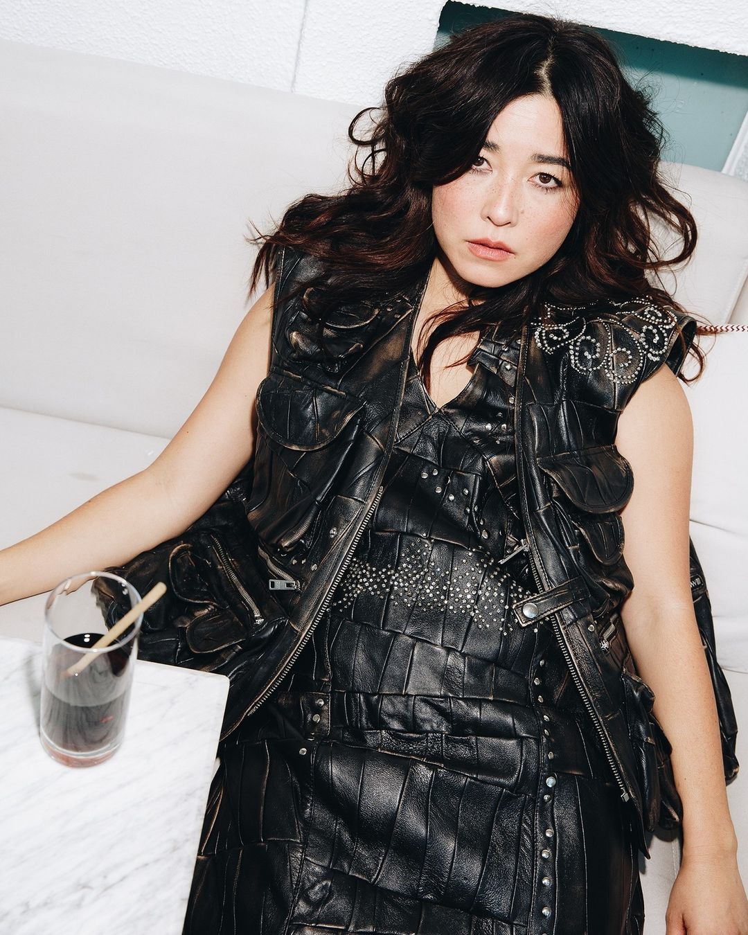 Portrait of an adult female wearing a black studded leather vest sitting indoors with a glass of dark liquid on a white marble table.