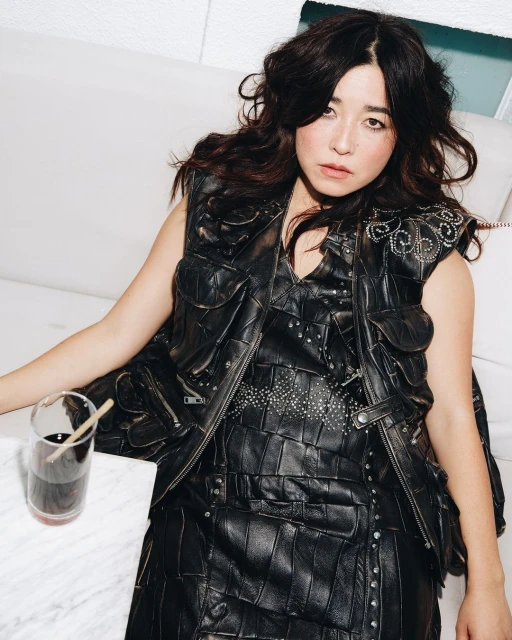 Portrait of an adult female wearing a black studded leather vest sitting indoors with a glass of dark liquid on a white marble table.