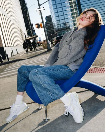 Young woman in a grey jacket and blue jeans seated on a bright blue curved chair outdoors on a sunny city sidewalk.