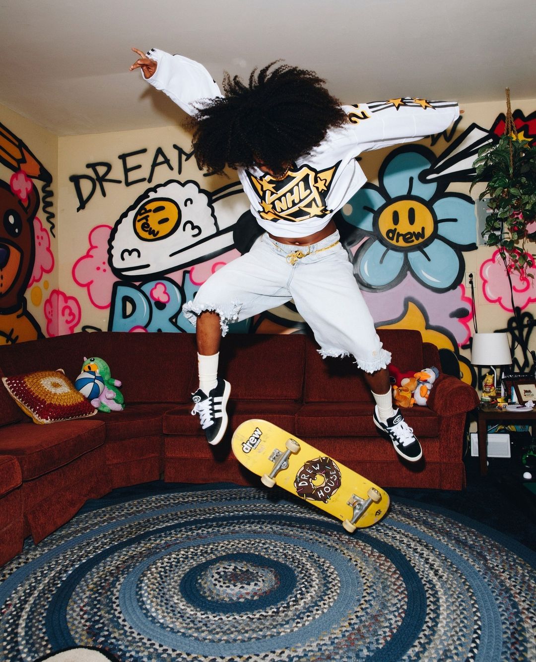 A young person with dark hair jumps with a yellow skateboard indoors, wearing a white top, light shorts, and sneakers, with graffiti walls.