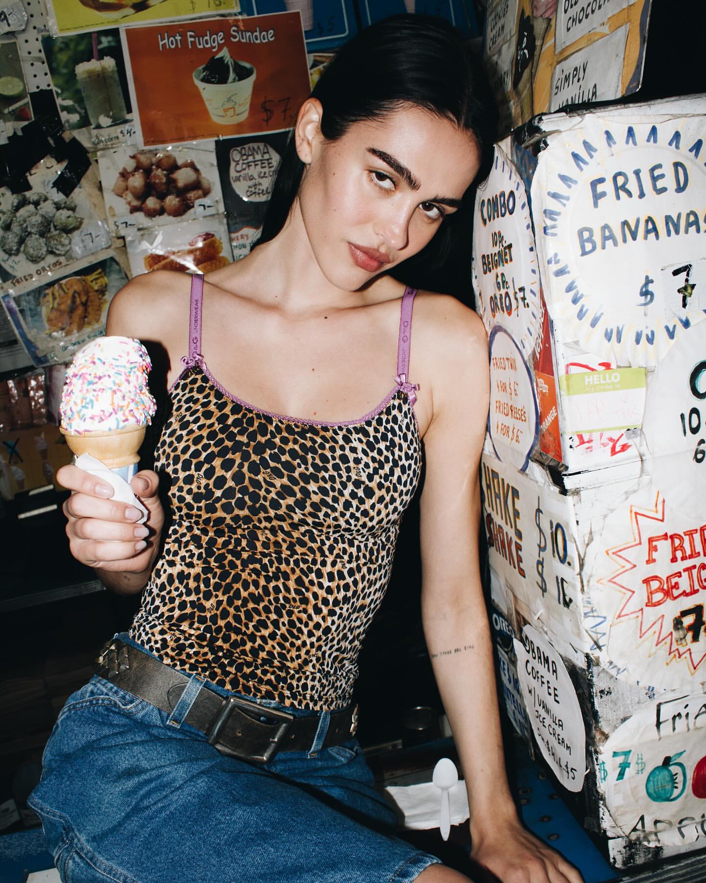 Brunette model in animal-print top, holds ice cream cone by vintage food stand with hand-lettered menus and retro vibes, eye-level.
