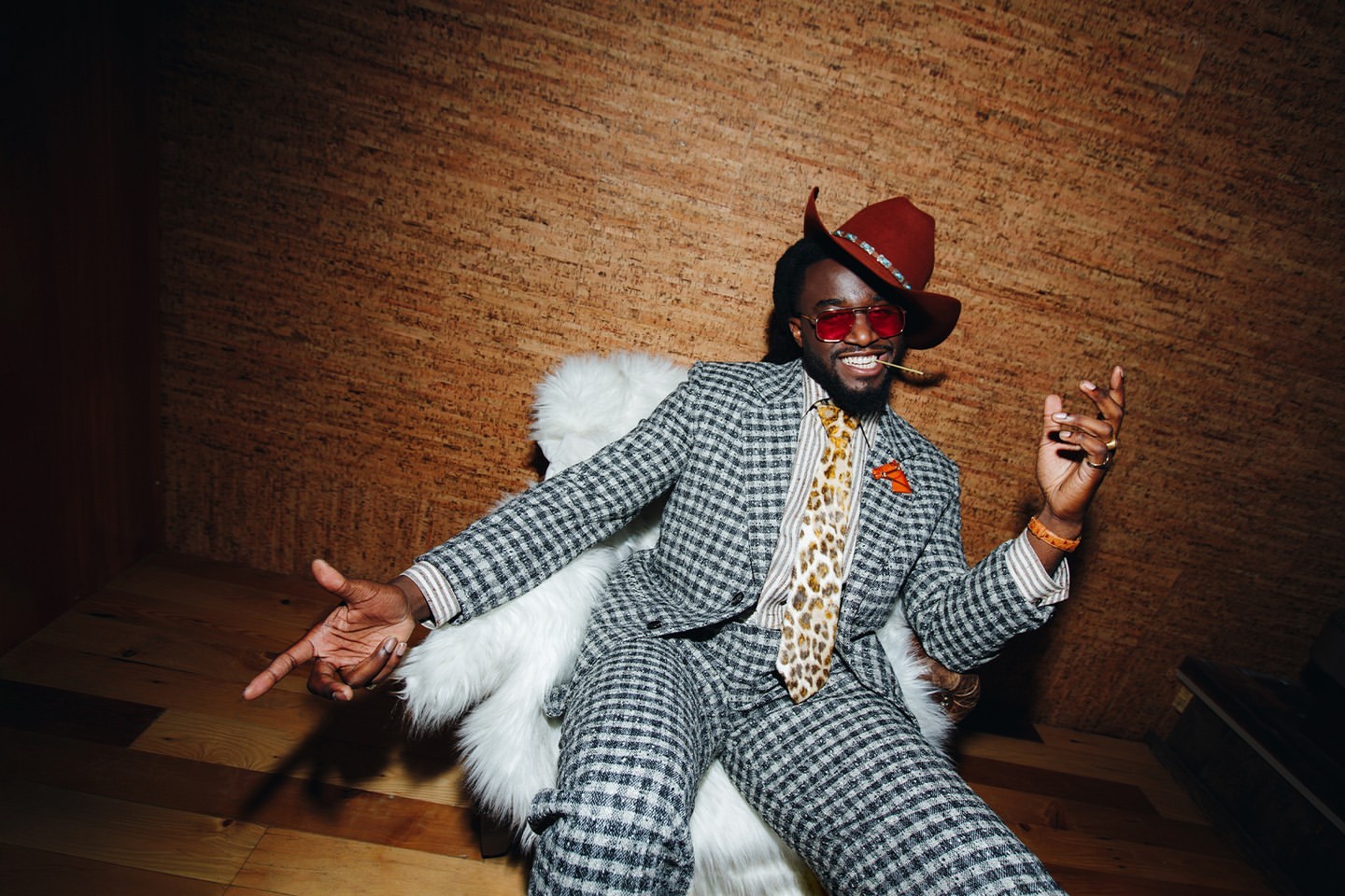 Stylish adult man with beard wearing a grey plaid suit, orange-tinted glasses, and a brown hat, posing on a white furry chair.