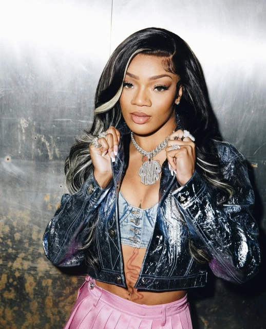 Close-up of a stylish young adult female wearing a metallic blue jacket, jewelry, and a pink skirt against a grey background.