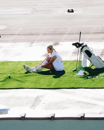 A woman in sportswear ties her shoe, seated on artificial grass, with a golf bag and practice balls nearby on a rooftop surface.