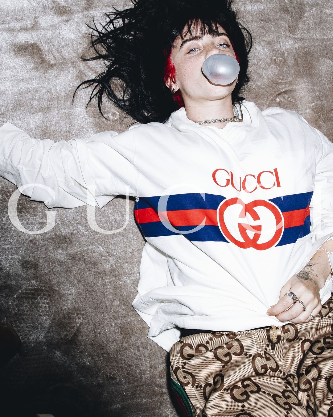 Young woman in a Gucci outfit blows a bubble while lying on a gray surface. Fashion editorial shot with retro styling and vibrant colors.
