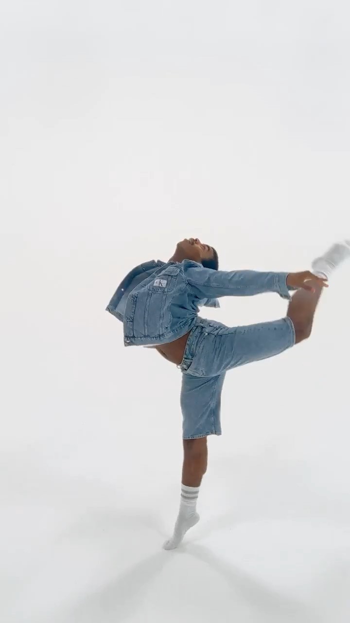 A black adult male in a denim jacket and shorts is balancing on one leg in a dance pose on a white background in a full shot.