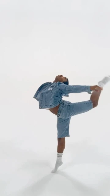 A black adult male in a denim jacket and shorts is balancing on one leg in a dance pose on a white background in a full shot.