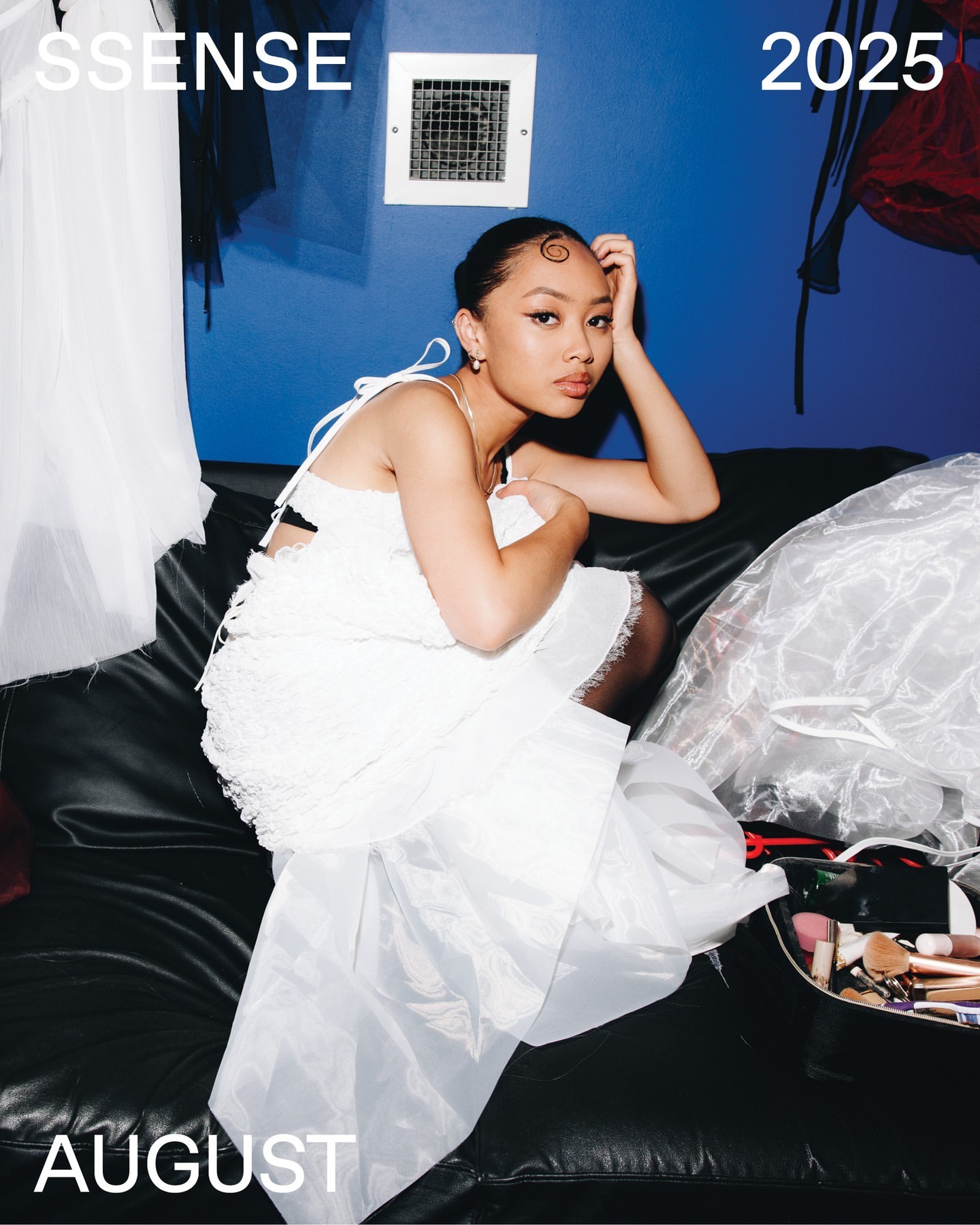A young adult woman wearing a white dress poses on a black couch in front of a blue wall with makeup kit and studio lighting.
