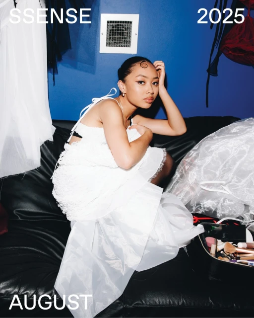 A young adult woman wearing a white dress poses on a black couch in front of a blue wall with makeup kit and studio lighting.