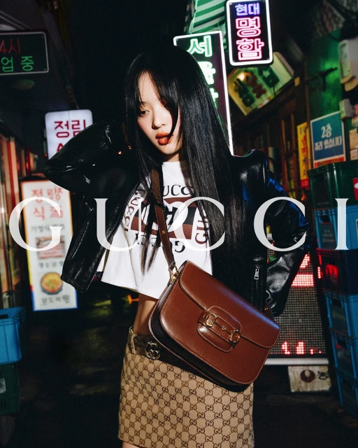 Young woman poses in an urban alley with a Gucci skirt, shirt, and handbag, illuminated by neon signs at night. Fashion editorial.