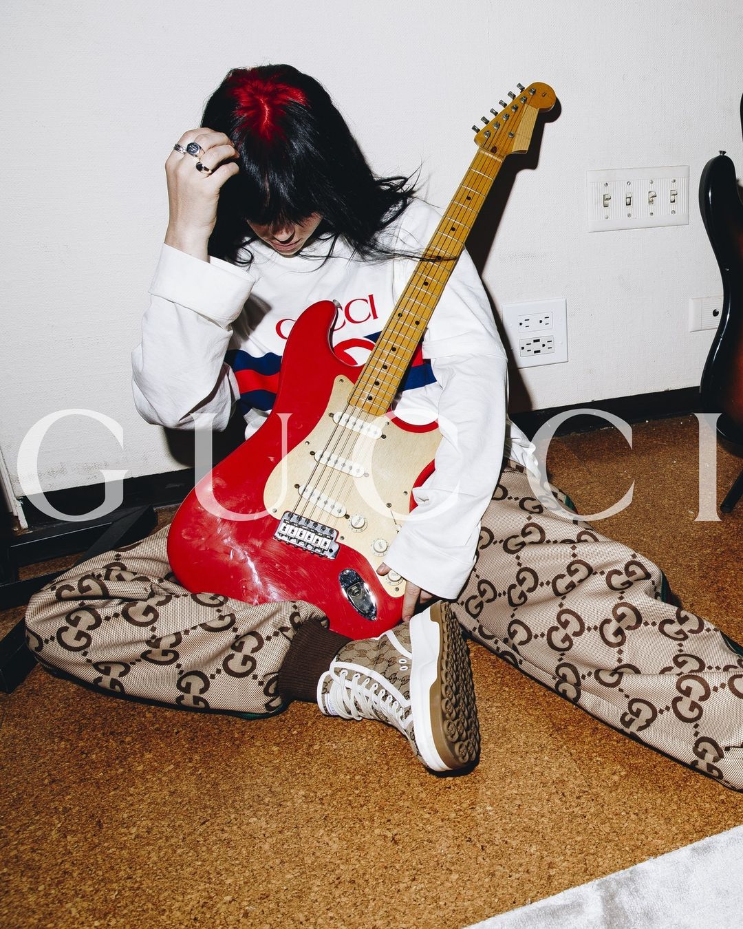 Young adult poses while sitting on the floor with an electric guitar, wearing Gucci brand printed apparel, long sleeve shirt and sneaker.
