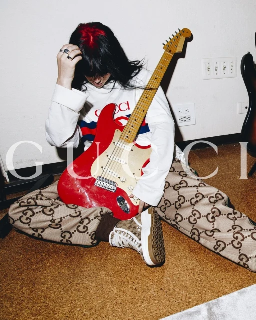 Young adult poses while sitting on the floor with an electric guitar, wearing Gucci brand printed apparel, long sleeve shirt and sneaker.