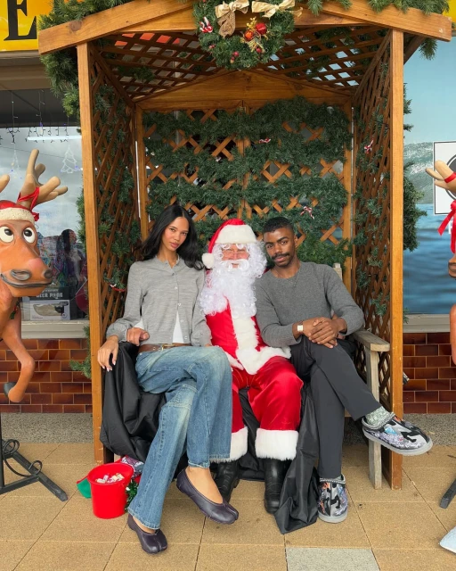 Two adults pose with Santa Claus on a bench decorated with greenery. A reindeer statue is in the foreground.