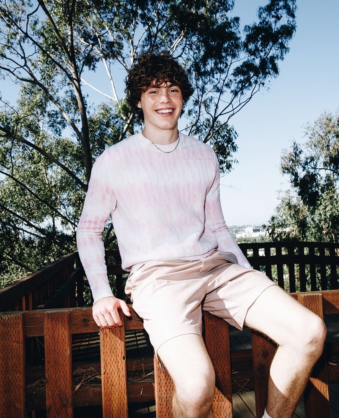 Young man smiles while sitting on a wooden fence outdoors. He wears a patterned long-sleeved shirt and shorts with trees in the background.