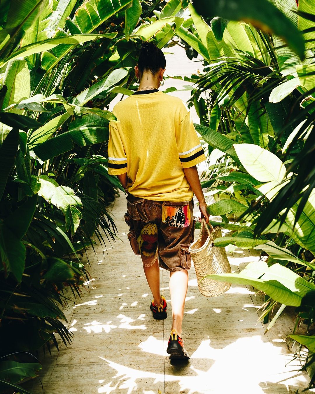 Woman walks away on a sunlit stone path through a tunnel of lush tropical green plants wearing a yellow t-shirt and cargo shorts.