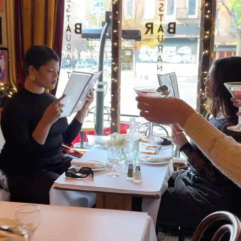 Two women enjoying drinks at a restaurant table, with one reading a menu and another toasting a martini in a bright indoor setting.