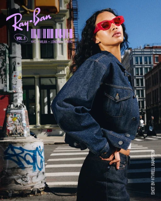 Woman in denim jacket and red sunglasses poses in an urban setting with buildings and a street, showcasing fashion and style.