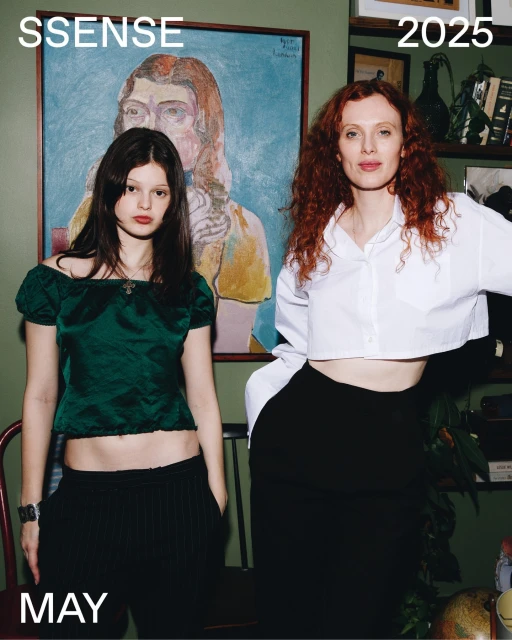 Two women pose indoors against an art-filled backdrop. One wears green, the other white and black, in a fashion editorial shot.
