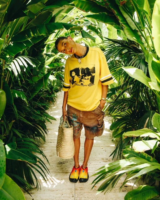 Woman in yellow t-shirt and brown shorts stands in a lush green outdoor setting on a light stone path, holding a woven bag.