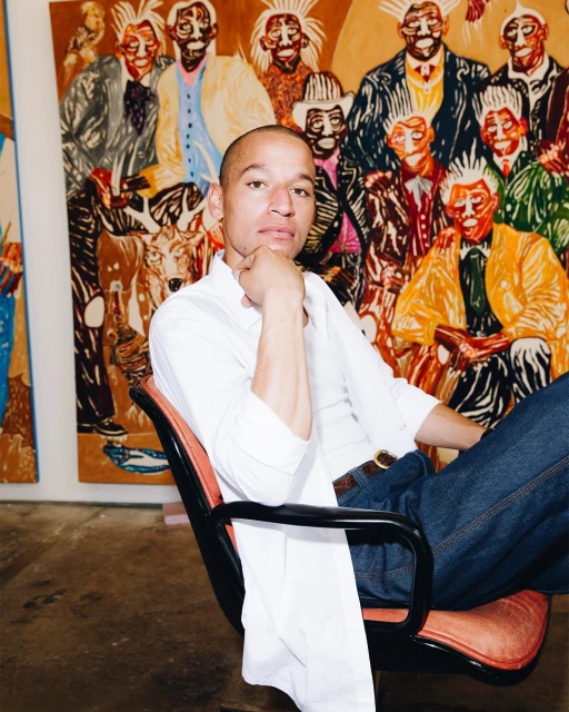 Adult male seated in a red chair with a pensive expression, paintings in the background in a well lit indoor setting.