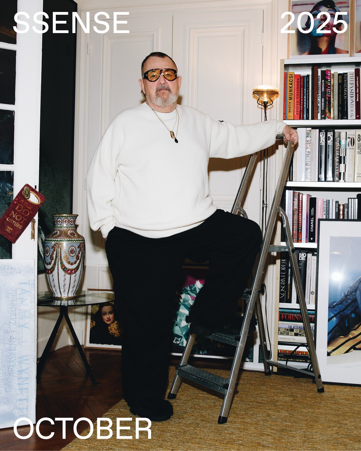Mature man in sunglasses poses with a ladder in an indoor setting with books and decorative items; cream, black and gold tones.