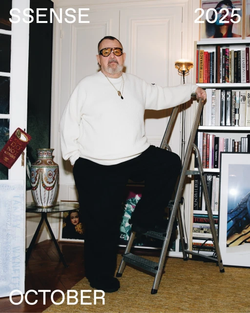 Mature man in sunglasses poses with a ladder in an indoor setting with books and decorative items; cream, black and gold tones.