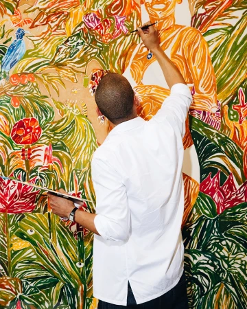 Artist in white shirt stands painting a colorful mural of people and plant life with a brush, viewed from behind in an indoor setting.