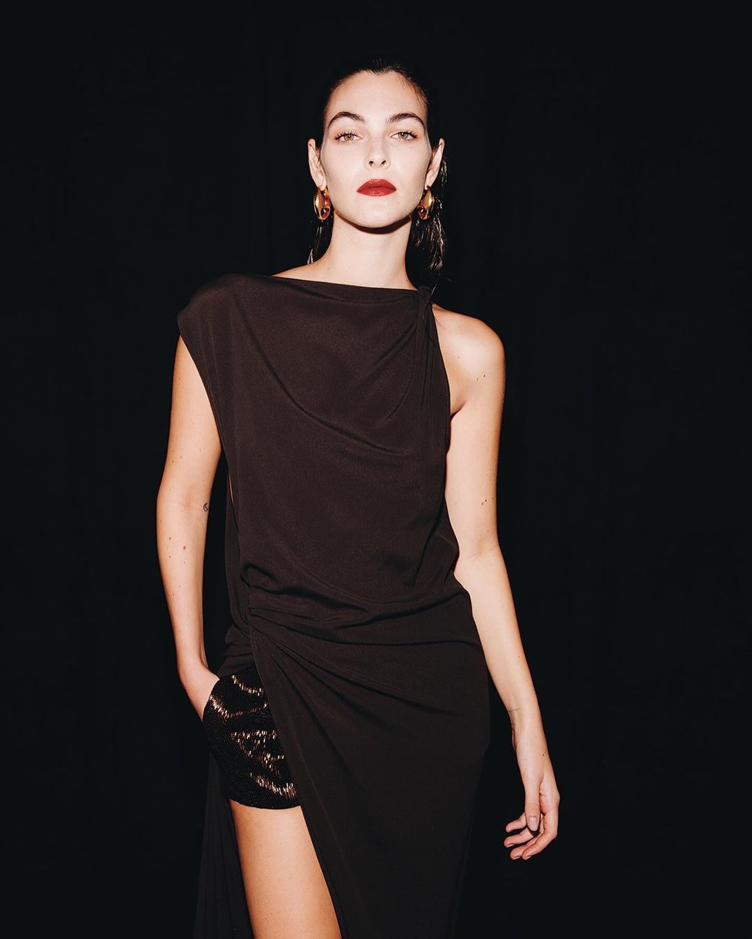 Woman in a black asymmetrical dress with dark hair and red lipstick against a black backdrop
