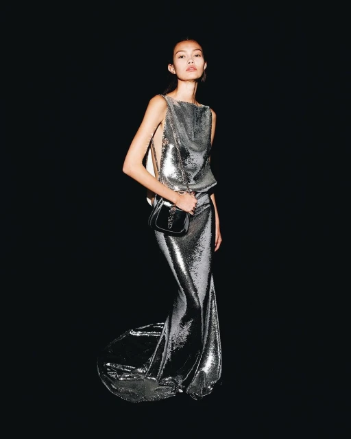 Elegant woman in a metallic silver gown and shoulder bag poses against a black backdrop, highlighting fashion and style.
