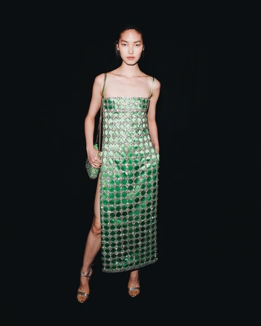 An Asian fashion model poses in a green sequin dress and strappy heels with a sequined purse against a plain black background.