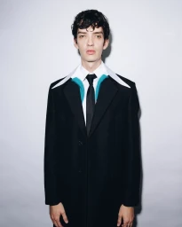 A man in a black coat and white collared shirt with blue accents, photographed by Tyrell Hampton against a plain background.