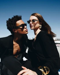 Two models wearing black apparel and sunglasses posing outdoors against a clear blue sky background