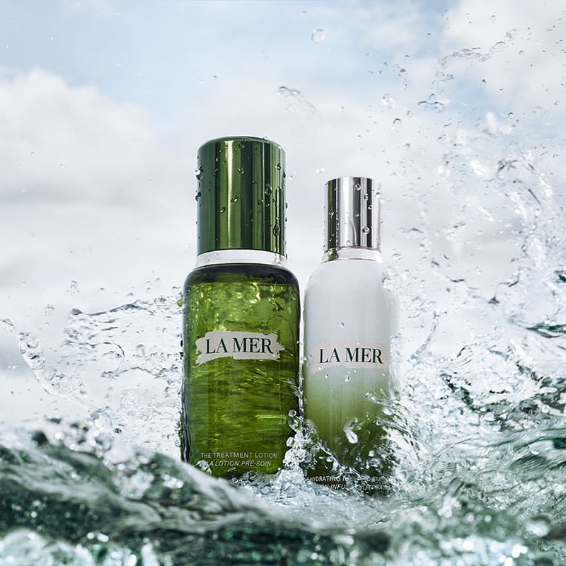 Two La Mer skincare bottles, one green and one white, surrounded by splashing water with a cloudy sky visible in the background.