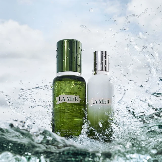 Two La Mer skincare bottles, one green and one white, surrounded by splashing water with a cloudy sky visible in the background.