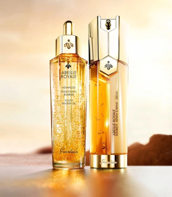 Two Guerlain Abeille Royale skincare products, including serum and watery oil, in gold packaging, set against a gradient background.