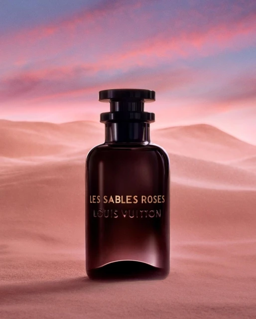 Luxury still life shot of a Les Sables Roses Louis Vuitton perfume bottle against a pink sand dune and sunset-colored sky background.