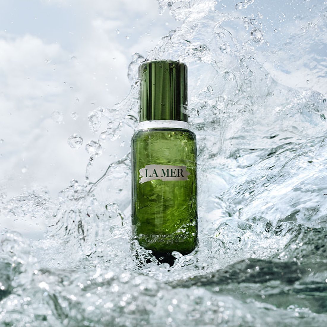 Green glass La Mer bottle surrounded by splashing water droplets against a light blue sky with clouds, product shot for skincare advertising.