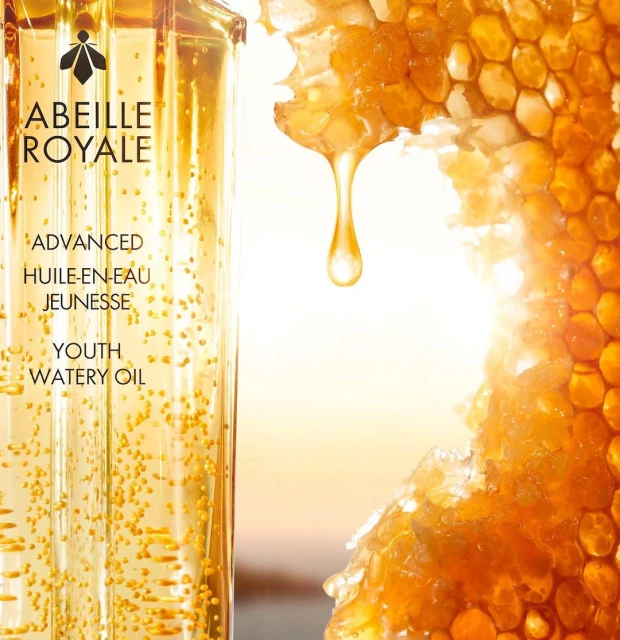 Product shot of Abeille Royale Youth Watery Oil, showcasing the golden liquid filled with micro-bubbles next to golden honeycomb with dripping honey.