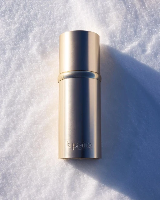 La Prairie cosmetic product in a gold container sits on snow with a long shadow. Beauty shot with a single object and no people.