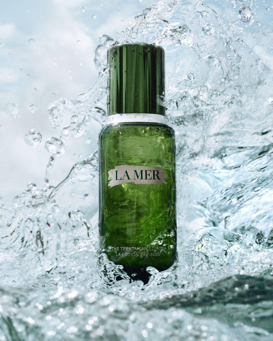 La Mer treatment lotion bottle, dark green with white lettering, surrounded by splashing water against a light blue sky background.