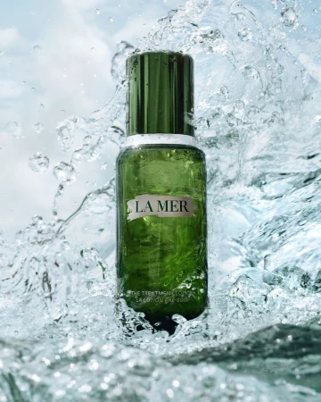 La Mer treatment lotion bottle, dark green with white lettering, surrounded by splashing water against a light blue sky background.