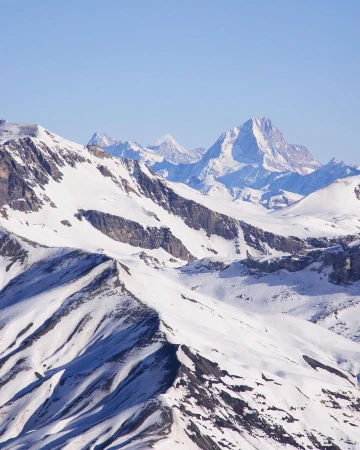 Snowy mountain range landscape under a clear blue sky. White snow covers the mountain peaks and slopes in the distance.