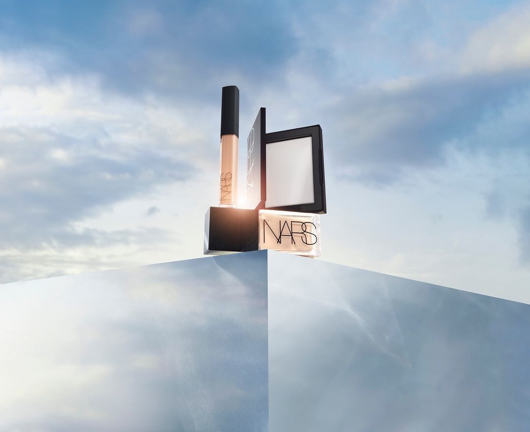 NARS makeup product arrangement against a cloudy sky background, featuring concealer, powder, and foundation. Beauty and cosmetic advertisement.