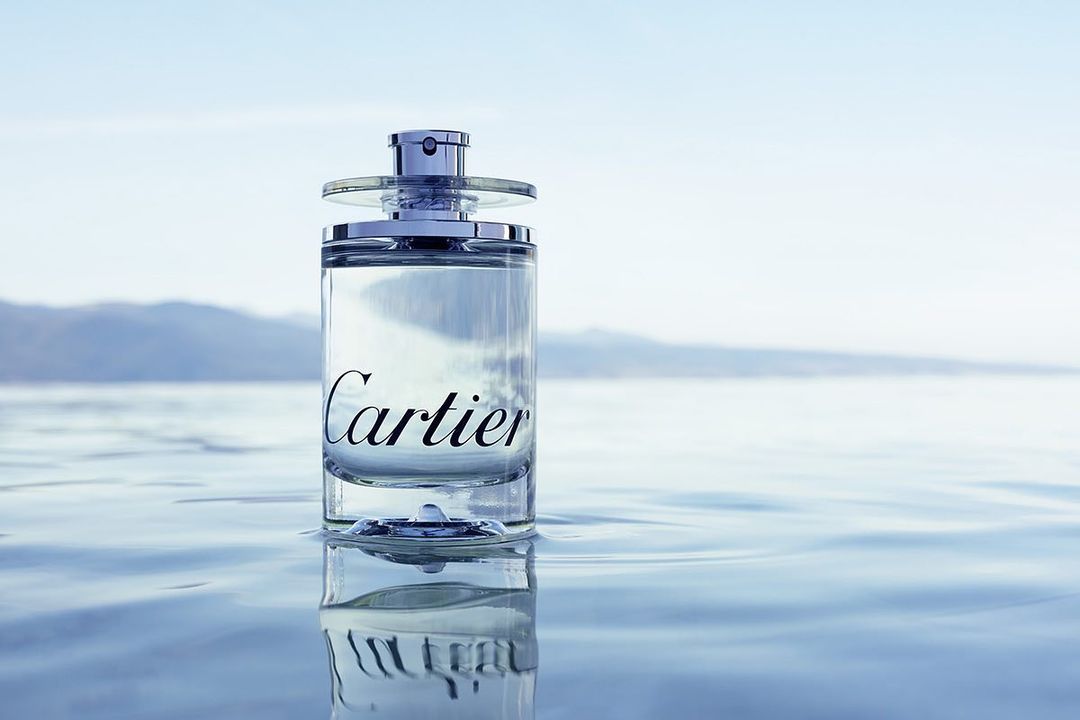 Glass Cartier perfume bottle with silver accents is sitting on top of blue water with mountains and light-blue sky in the distance.